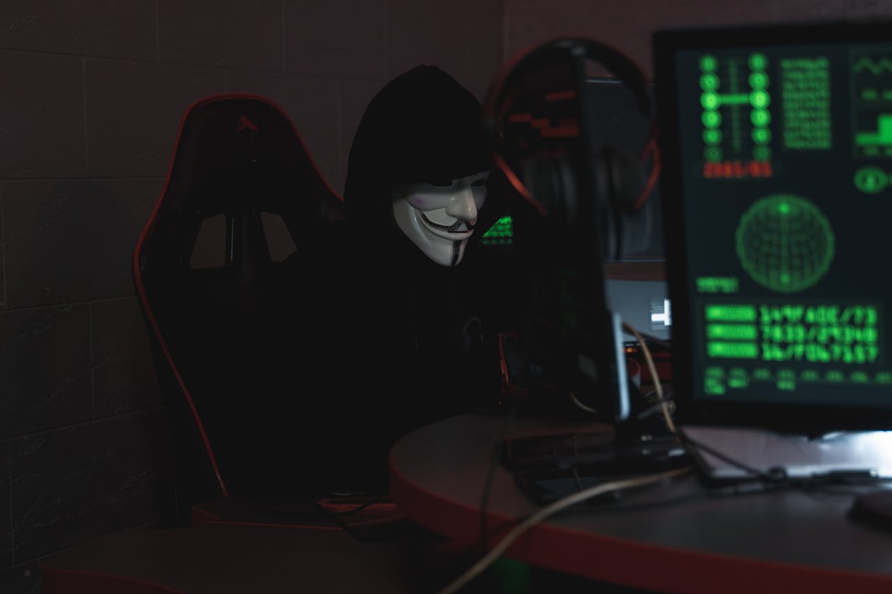 An anonymous hacker wearing a Guy Fawkes mask sits at a computer in a dimly lit room, engaged in cyber activities.