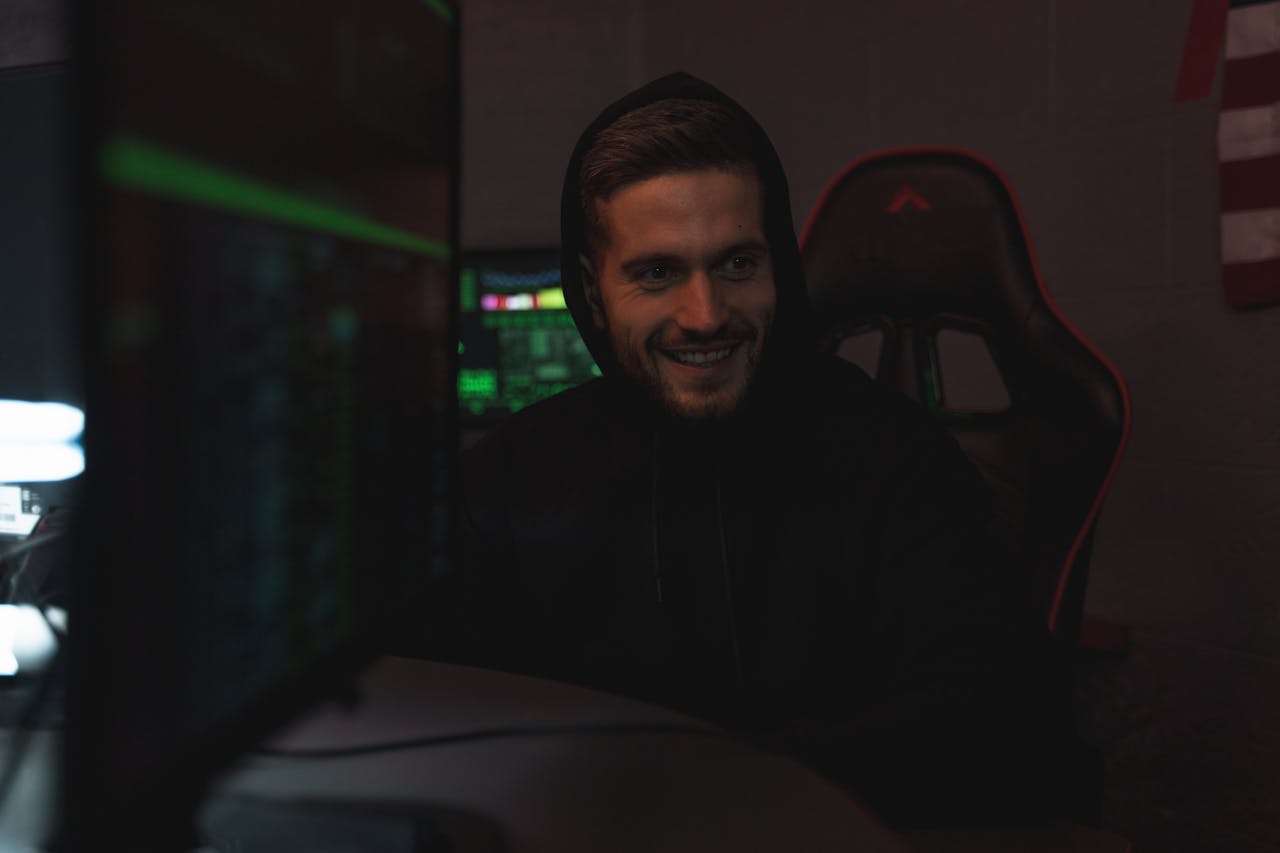 menu-07 Man in a black hoodie sits in a dark room, smiling while using a computer. Tech-related setting.