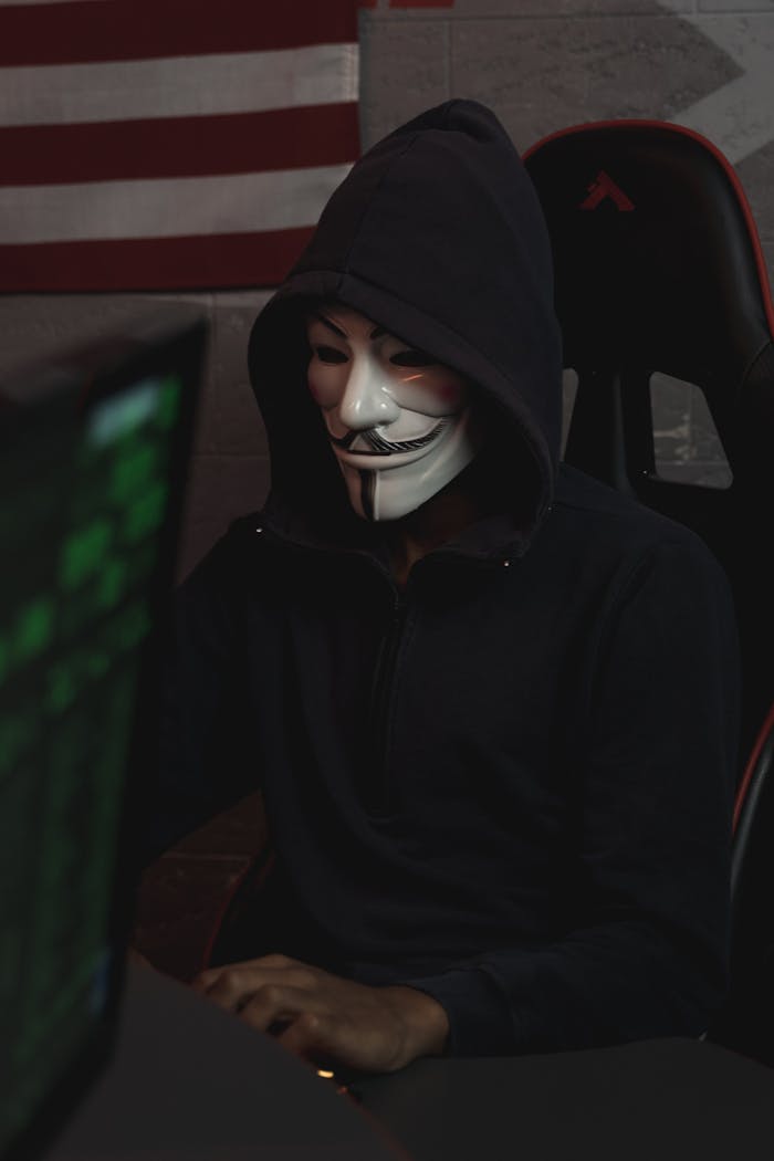menu-17 Masked hacker wearing a Guy Fawkes mask operates a computer, symbolizing cyber security threats.