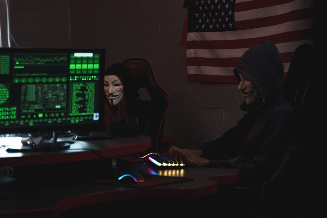 about-02 Two anonymous individuals wearing hacker masks use computers in a dark room with an American flag backdrop, highlighting cyber security themes.