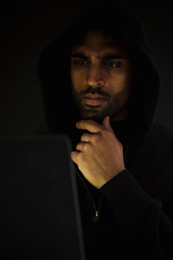 menu-02 Concentrated bearded male hacker wearing black hood looking at netbook screen and touching chin in contemplation while hacking software in darkness